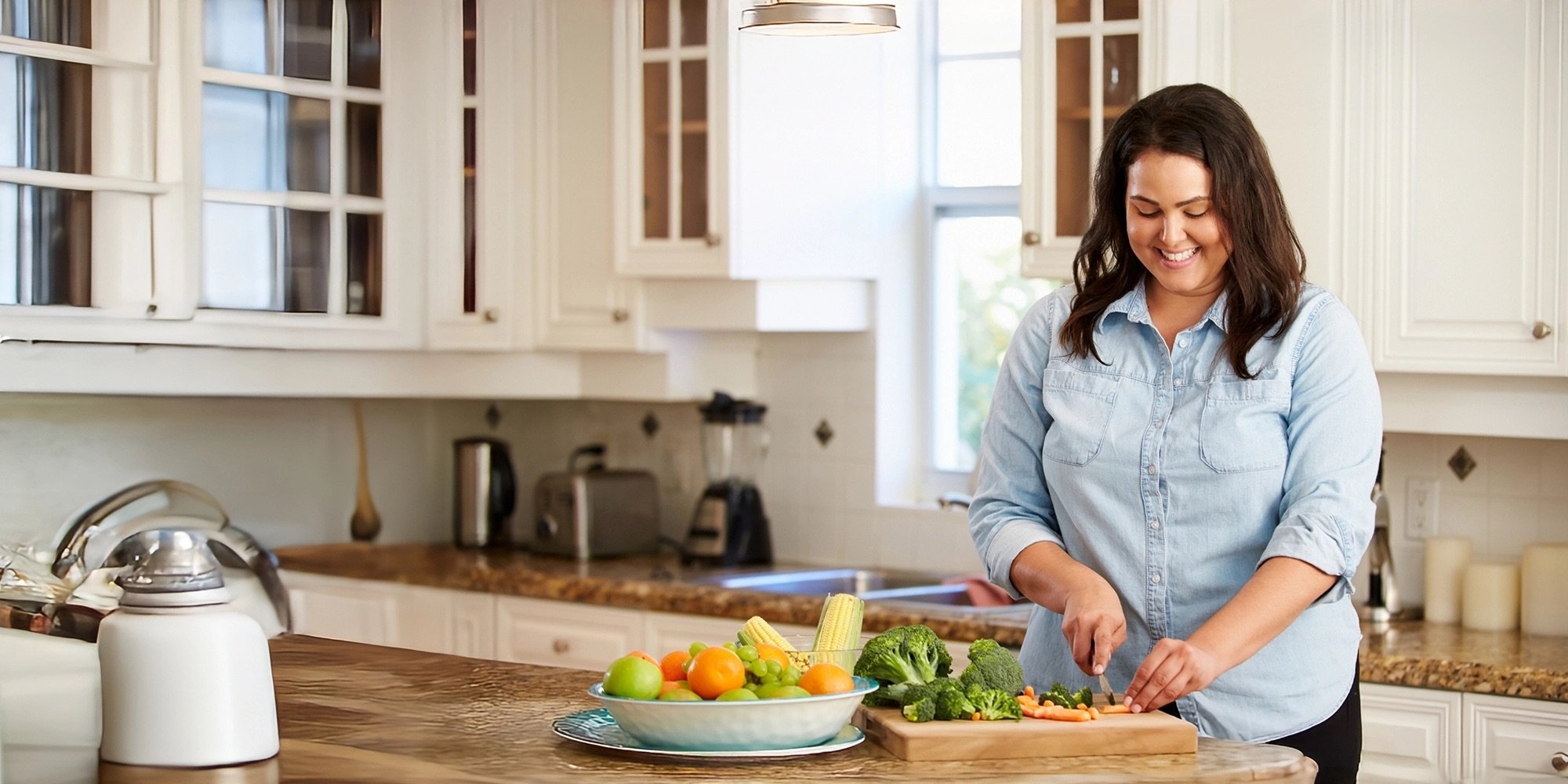 A smiling woman stands in her kitchen, cutting vegetables at the counter